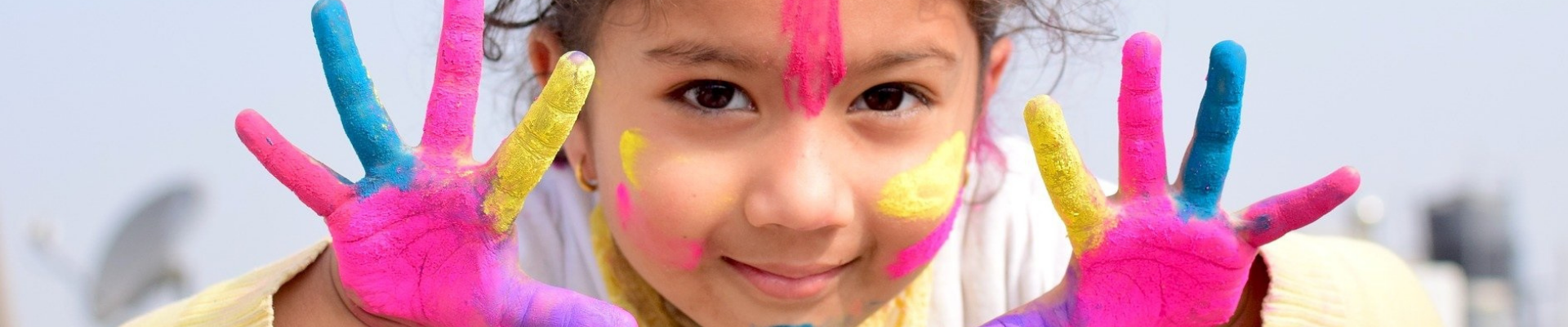 Ein Kind hält seine Hände in die Kamera, die mit gelber, blauer und rosaner Farbe bemalt sind. Auch im Gesicht hat das Kind bunte Farbe.