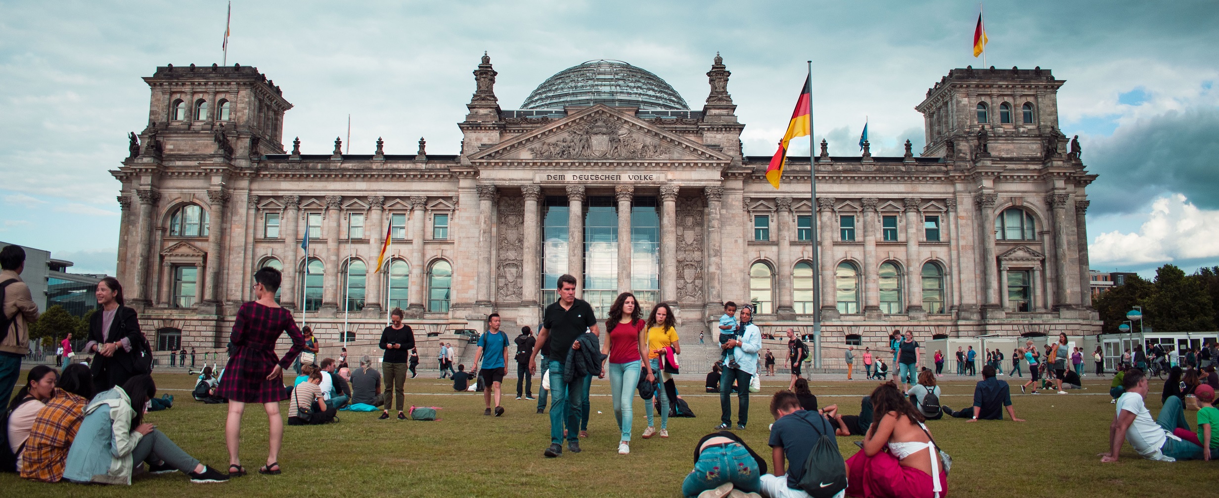 Zu sehen ist der Bundestag. Davor sitzen Menschen auf der Wiese.