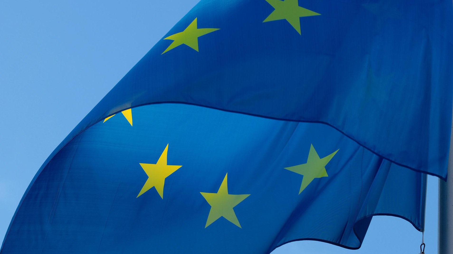 Zu sehen ist die europäische Flagge, welche im Kreis angeordnete Sterne zeigt und einen blauen Hintergrund hat.