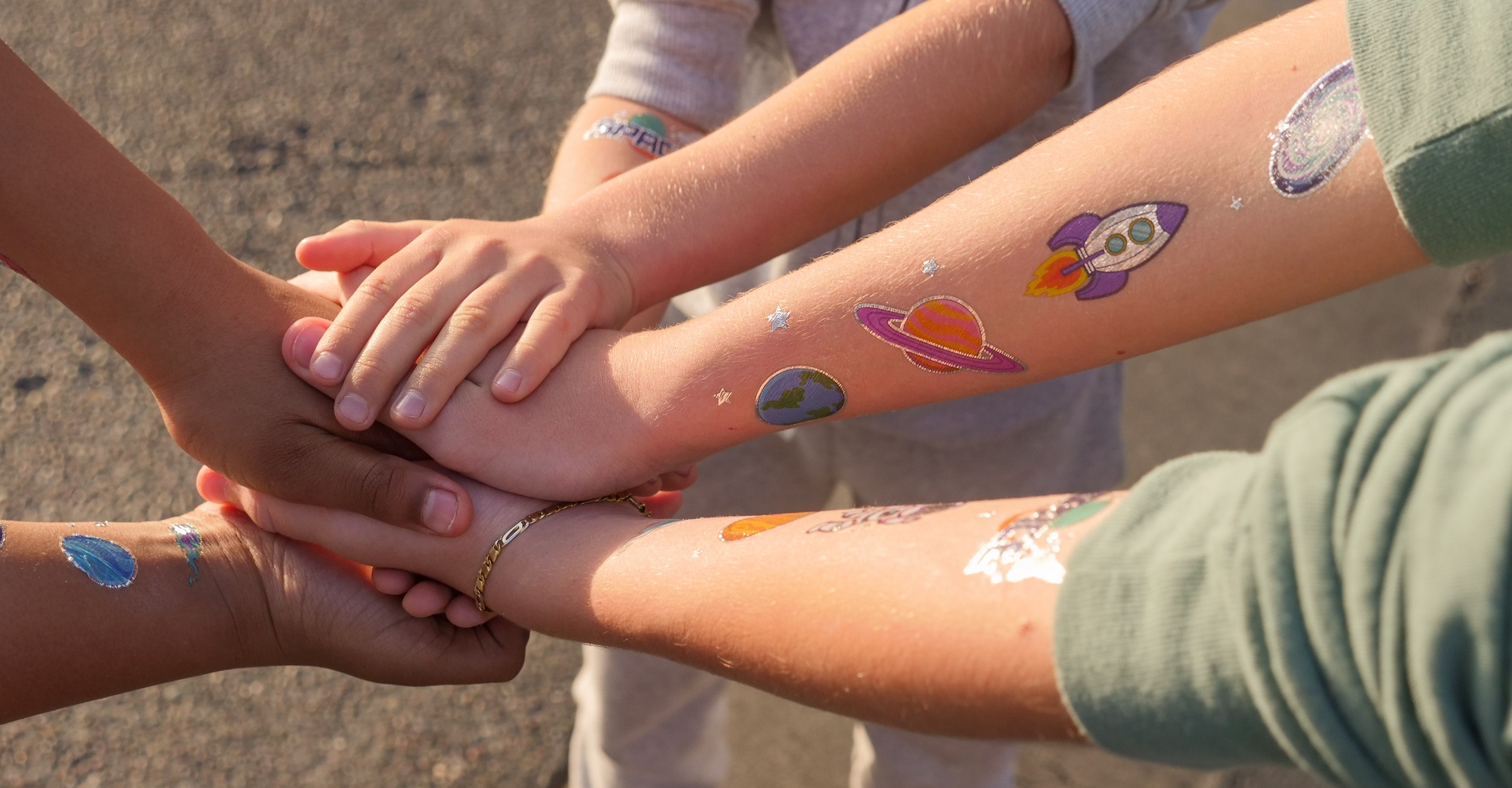 Junge Menschen legen die Hände aufeinander und halten zusammen. Auf den Armen sind bunte Tattoos von Planeten aufgeklebt.