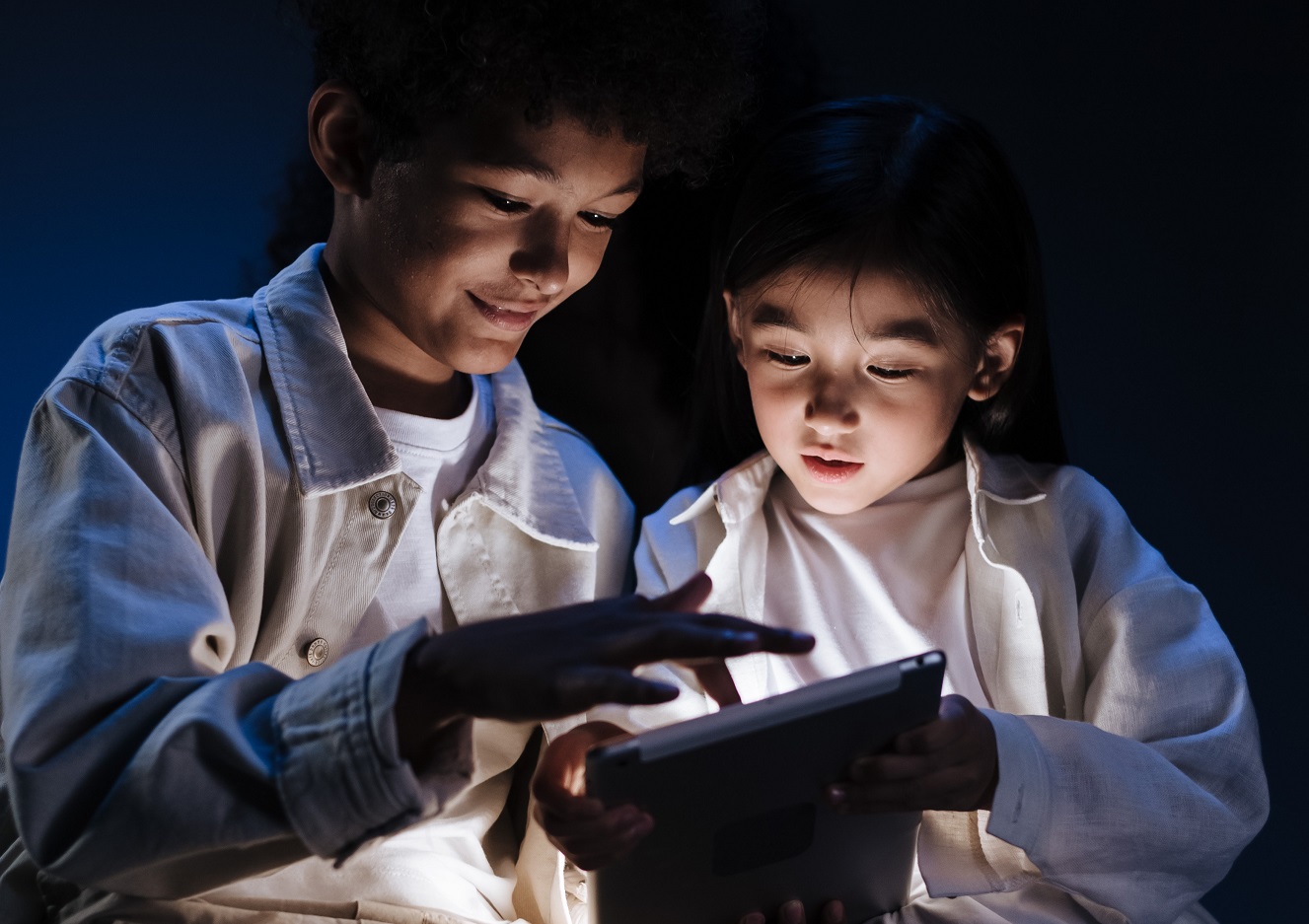 Zwei Kinder sehen auf ein I-Pad, welches ell im Dunkeln leuchtet.
