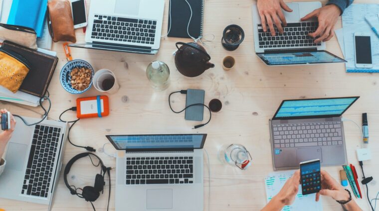 Perspektive von Oben, mehrere Laptops stehen auf einem Tisch, Menschen arbeiten daran, daneben liegen Snacks und Kaffeetassen