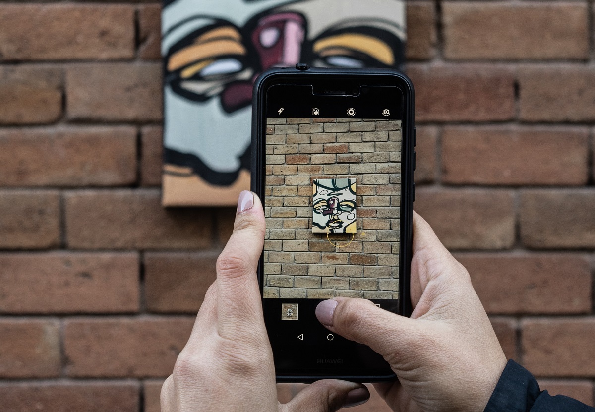 Nahaufnahme von einer Person, die mit einem Smartphone ein Gemälde fotografiert, dass an einer Steinmauer hängt